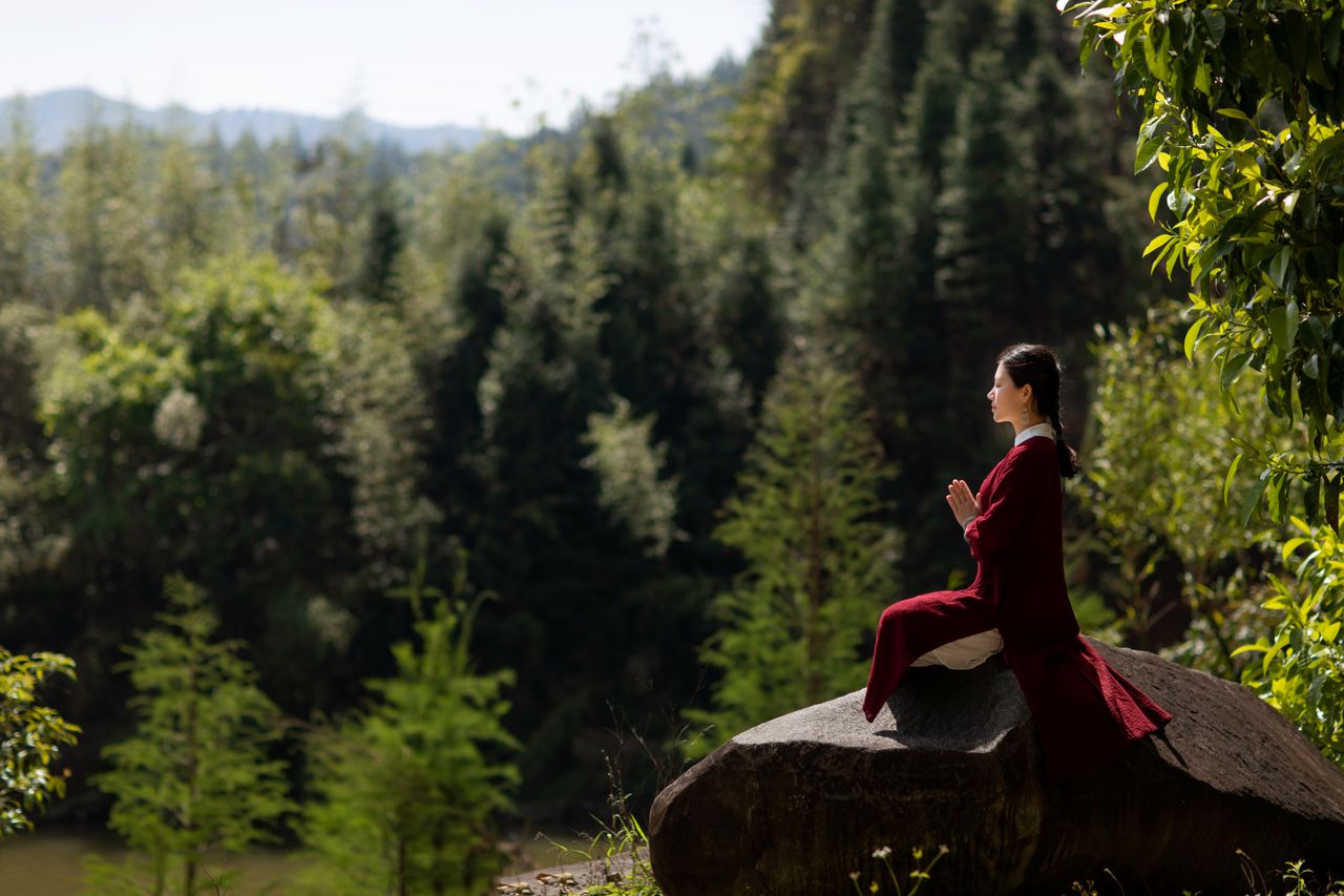 Asian woman in traditional clothing meditating on a rock in a serene forest setting.